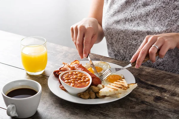 Žena jíst tradiční anglickou snídani smažená vejce, párky, fazole, houby, Grilovaná rajčata, slanina, káva a pomerančový džus, na dřevěný stůl Stock Obrázky