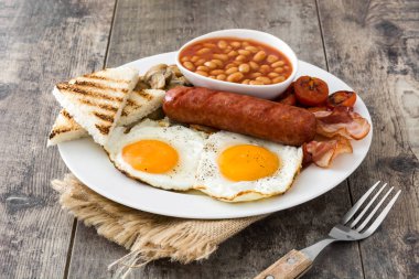 Geleneksel İngiliz kahvaltısı kızarmış yumurta, sosis, fasulye, mantar, ızgara domates ve ahşap arka planda pastırma.