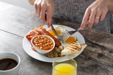 Geleneksel bir İngiliz kahvaltısı ile kızarmış yumurta, sosis, fasulye, mantar, ızgara domates, pastırma, kahve ve portakal suyu, tahta masada yemek kadın