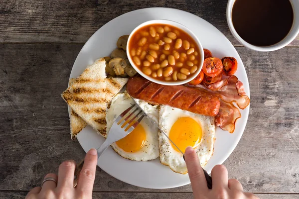 Mulher comendo um café da manhã tradicional inglês com ovos fritos, salsichas, feijão, cogumelos, tomates grelhados, bacon, café e suco de laranja, na mesa de madeira — Fotografia de Stock