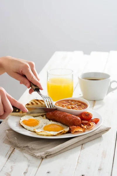 Mulher comendo tradicional completo Inglês café da manhã com ovos fritos, salsichas, feijão, cogumelos, tomates grelhados e bacon no fundo de madeira branco — Fotografia de Stock