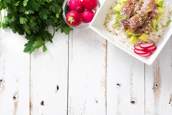 Hawaiian tuna poke bowl with avocado, radishes and sesame seeds on white wooden background