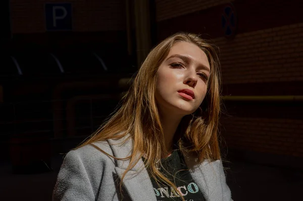 Elegante chica bonita joven con un abrigo gris mira a la cámara y sonríe ligeramente contra el fondo de la ciudad . — Foto de Stock