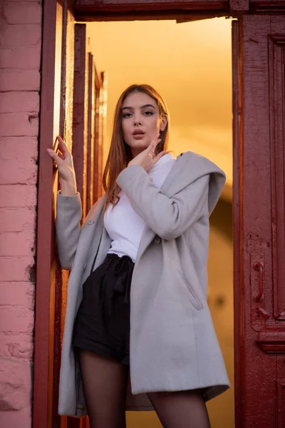 Menina bonita nova elegante em um casaco cinza olha para a câmera e sorri ligeiramente contra o fundo da cidade . — Fotografia de Stock