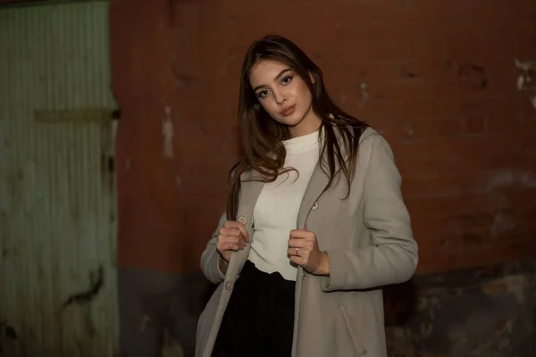 Elegante chica bonita joven con un abrigo gris mira a la cámara y sonríe ligeramente contra el fondo de la ciudad . —  Fotos de Stock