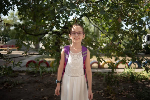 Skolflicka med en violett ryggsäck och lila glasögon. Flicka i vit klänning, glasögon på hösten. — Stockfoto