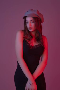 Göz kamaştırıcı genç kadın, gri duvarda dalgalı saçlarla poz veren şık elbiseler giyiyor.