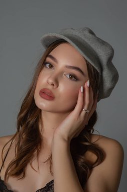 Göz kamaştırıcı genç kadın, gri duvarda dalgalı saçlarla poz veren şık elbiseler giyiyor.