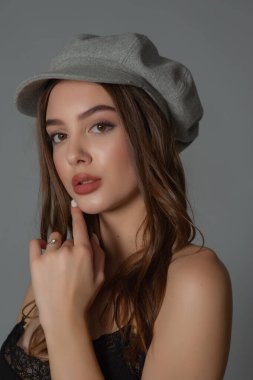 Göz kamaştırıcı genç kadın, gri duvarda dalgalı saçlarla poz veren şık elbiseler giyiyor.