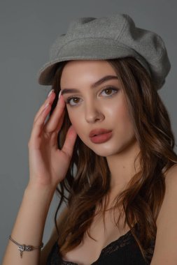 Göz kamaştırıcı genç kadın, gri duvarda dalgalı saçlarla poz veren şık elbiseler giyiyor.