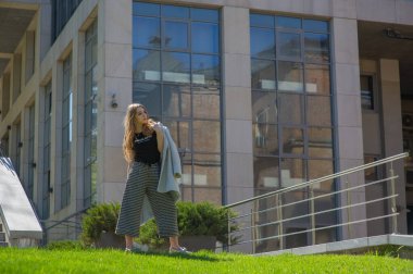 Göz kamaştırıcı genç kadın, gri duvarda dalgalı saçlarla poz veren şık elbiseler giyiyor.