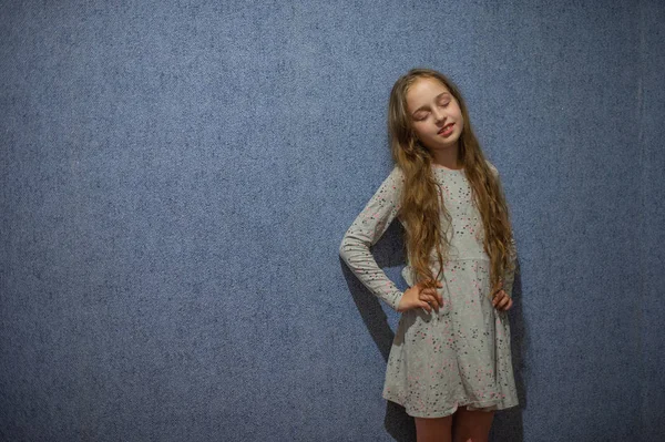 Söt flicka elva år med blont långt hår i en grå klänning på en blå bakgrund — Stockfoto
