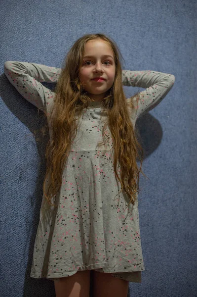 Schattig meisje van elf jaar oud met blond lang haar in een grijze jurk op een blauwe achtergrond — Stockfoto