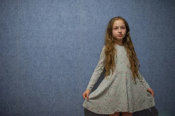 Mignonne fille de onze ans avec des cheveux longs blonds dans une robe grise sur un fond bleu — Photo