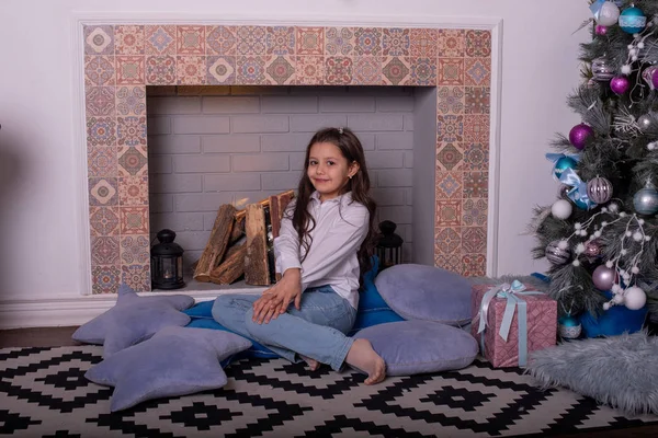Dziewczyna w dżinsach siedzi na białej kanapie w pobliżu dekoracji Nowego Roku. Dziewczyna w dżinsach w pobliżu noworocznego kominka — Zdjęcie stockowe