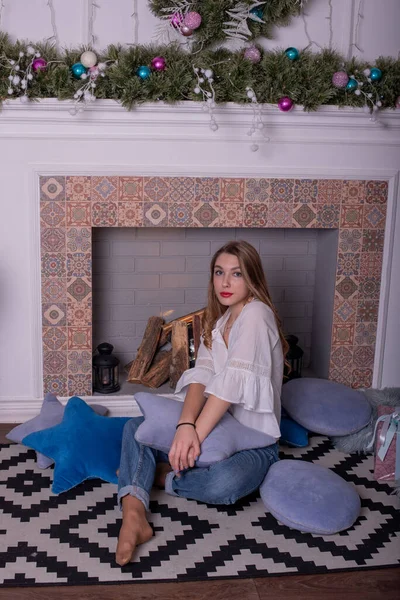 Dziewczyna w dżinsach siedzi na białej kanapie w pobliżu dekoracji Nowego Roku. Dziewczyna w dżinsach w pobliżu noworocznego kominka — Zdjęcie stockowe