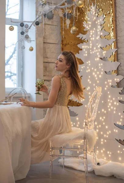 Mädchen in einem glänzenden Abendkleid. Heiligabend. Gemütlicher Urlaub an der Tanne mit Lichtern und Golddekor — Stockfoto