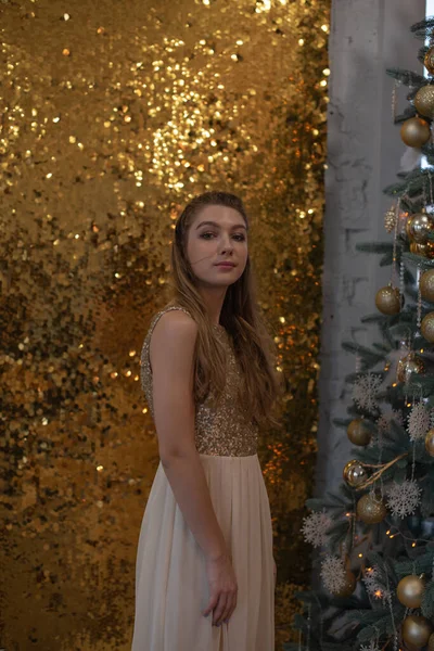 Parlak bir gece elbisesi giyen kız. Noel arifesi. Kürk ağacında rahat bir tatil Işıklar ve altın dekorla — Stok fotoğraf