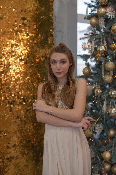 Chica en un vestido de noche brillante. Nochebuena. Acogedoras vacaciones en el árbol de piel con luces y decoración dorada —  Fotos de Stock