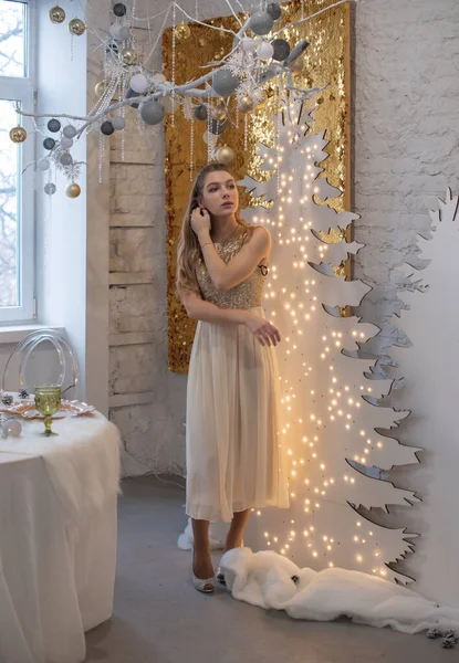 girl in a shiny evening dress. Christmas Eve. Cozy holiday at the fur-tree with lights and gold decor