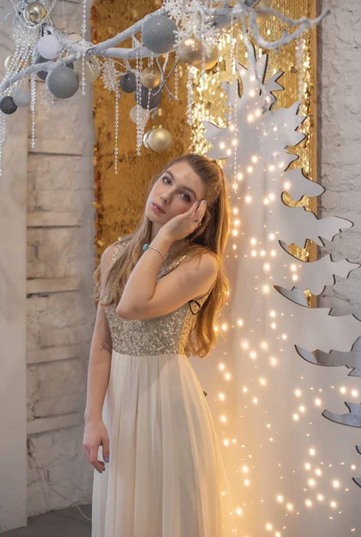 Meisje in een glanzende avondjurk. Kerstavond. Gezellige vakantie aan de pelsboom met lichtjes en gouden decor — Stockfoto