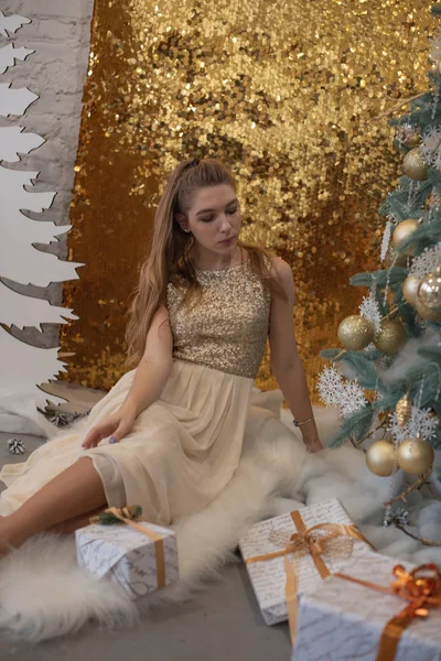 Chica en un vestido de noche brillante. Nochebuena. Acogedoras vacaciones en el árbol de piel con luces y decoración dorada —  Fotos de Stock