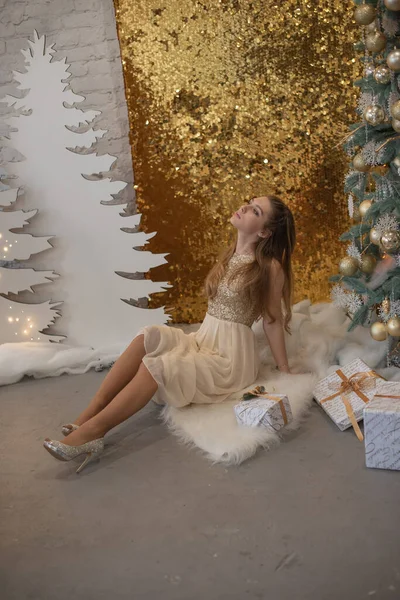Menina em um vestido de noite brilhante. Véspera de Natal. Férias acolhedoras na árvore de peles com luzes e decoração dourada — Fotografia de Stock