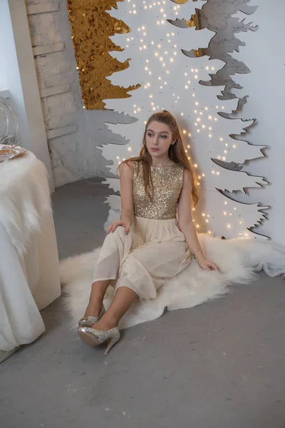 Chica en un vestido de noche brillante. Nochebuena. Acogedoras vacaciones en el árbol de piel con luces y decoración dorada —  Fotos de Stock