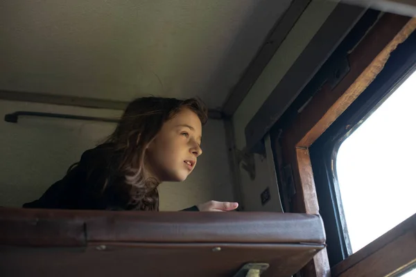 Linda chica acostada en el estante superior en el tren y mirando por la ventana a la calle. Vacaciones de verano — Foto de Stock