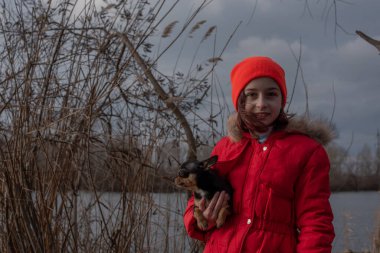 Hanımefendinin ceketinin altında küçük bir chihuahua köpeği ısıtılıyor. 9 yaşındaki bir kız nehir kenarında ceketli.