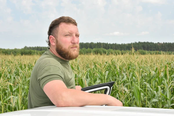 Fermier lângă câmpul de porumb în timpul verii. Un om pe fundalul unui câmp cu o mașină de porumb albă — Fotografie, imagine de stoc