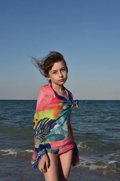 Chica hermosa y delgada en una túnica voladora de verano camina a lo largo de la costa del mar al atardecer. tarjeta de verano, espacio para copiar. Bebé en el mar. Viento en el pelo de una chica. Vacaciones de verano junto al mar — Foto de Stock