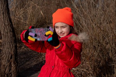 Sevimli kız, kırmızı ceketli, fotoğraf çekmek için poz veriyor ya da akıllı telefonuyla kendi fotoğrafını çekiyor.