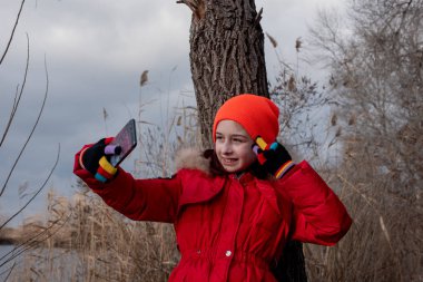 Sevimli kız, kırmızı ceketli, fotoğraf çekmek için poz veriyor ya da akıllı telefonuyla kendi fotoğrafını çekiyor.