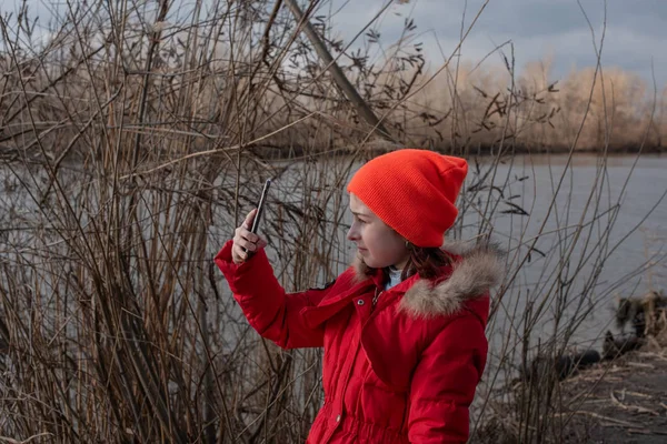 Gadis manis, mengenakan jaket merah, berpose untuk membuat selfie atau foto dirinya dengan smartphone — Stok Foto