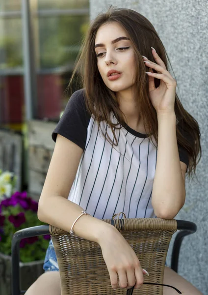 Mujer hipster joven bastante sexy en una camiseta i en una silla de paja al aire libre en un café en un día de verano. Atractiva morena — Foto de Stock