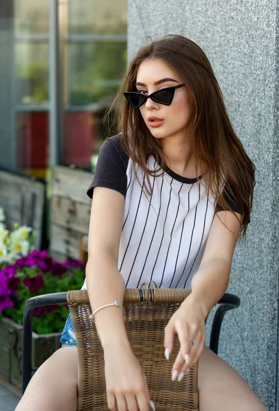 Pretty sexy young hipster woman in a T-shirt i on a straw chair outdoors in a cafe on a summer day. Attractive brunette — 스톡 사진