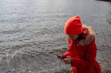 Sevimli kız, kırmızı ceketli, fotoğraf çekmek için poz veriyor ya da akıllı telefonuyla kendi fotoğrafını çekiyor.