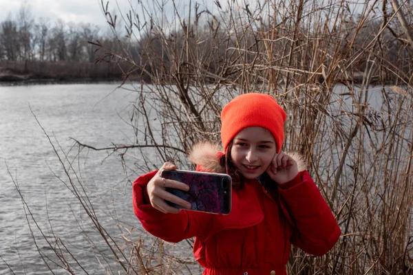 Adorable Girl, ubrana w czerwoną kurtkę, pozuje do robienia selfie lub zdjęcie siebie ze smartfonem — Zdjęcie stockowe