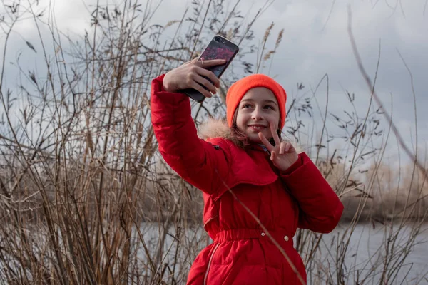 Adorable Girl, ubrana w czerwoną kurtkę, pozuje do robienia selfie lub zdjęcie siebie ze smartfonem — Zdjęcie stockowe