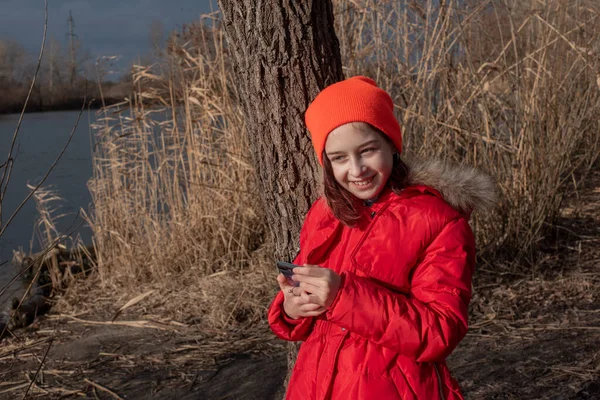 Adorable Girl, ubrana w czerwoną kurtkę, pozuje do robienia selfie lub zdjęcie siebie ze smartfonem — Zdjęcie stockowe