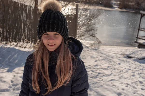 在寒冷的季节里 一个戴着帽子的漂亮金发姑娘的肖像展现了城市的风貌 走在街上的女孩 冬天穿黑色帽子和蓝色羽绒服的女孩 9岁女孩 — 图库照片
