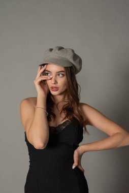 Göz kamaştırıcı genç kadın gri duvarda dalgalı saçlarıyla şık bir elbise giyiyor. Siyah elbiseli ve gri şapkalı bir kız elini öne uzattı. Kahverengi kıvırcık saçlı güzel bir genç kız. portre