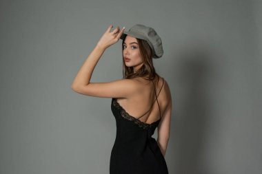 Göz kamaştırıcı genç kadın gri duvarda dalgalı saçlarıyla şık bir elbise giyiyor. Siyah elbiseli ve gri şapkalı bir kız elini öne uzattı. Kahverengi kıvırcık saçlı güzel bir genç kız. portre