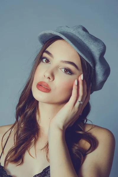 Glamorous young woman wears fashionable dress posing with fluttering hair at grey wall. Girl put forward hand in a black dress and a gray cap. Beautiful young girl with brown curly hairs. portrait