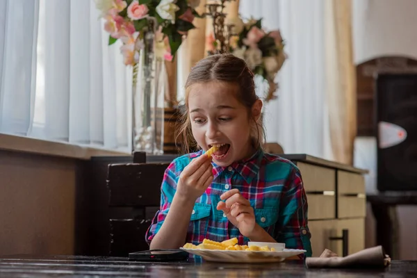 girl eating french-fries. Girl eating french fries in a cafe. A girl of 9 years old has lunch with french fries. French fries, sauce, lunch, food. A schoolgirl is eating and looking at a smartphone.