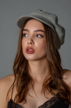 Göz kamaştırıcı genç kadın gri duvarda dalgalı saçlarıyla şık bir elbise giyiyor. Siyah elbiseli ve gri şapkalı bir kız elini öne uzattı. Kahverengi kıvırcık saçlı güzel bir genç kız. portre