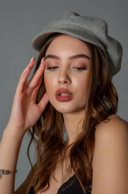 Göz kamaştırıcı genç kadın gri duvarda dalgalı saçlarıyla şık bir elbise giyiyor. Siyah elbiseli ve gri şapkalı bir kız elini öne uzattı. Kahverengi kıvırcık saçlı güzel bir genç kız. portre