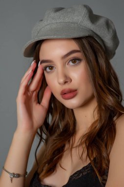 Göz kamaştırıcı genç kadın gri duvarda dalgalı saçlarıyla şık bir elbise giyiyor. Siyah elbiseli ve gri şapkalı bir kız elini öne uzattı. Kahverengi kıvırcık saçlı güzel bir genç kız. portre