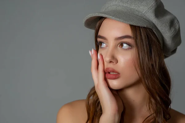 Glamorous young woman wears fashionable dress posing with fluttering hair at grey wall. Girl put forward hand in a black dress and a gray cap. Beautiful young girl with brown curly hairs. portrait